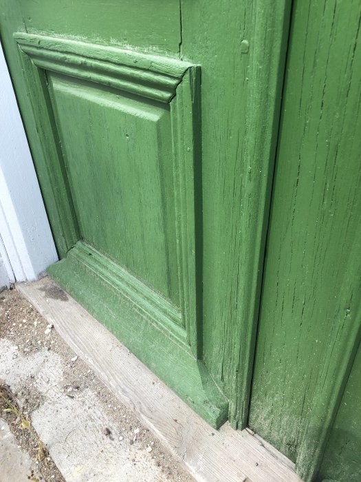Nedre delen av en grön trädörr med dekorativa detaljer och tecken på slitage.