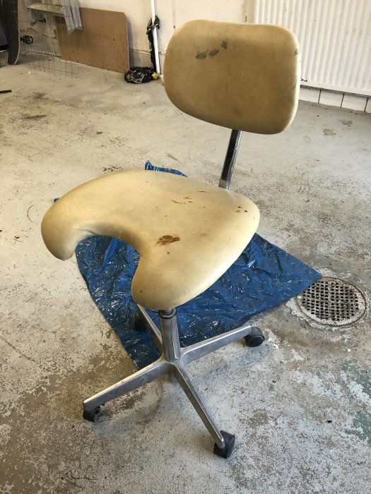 En sliten tandläkarstol med beige sits och synliga fläckar står i ett garage med betonggolv.