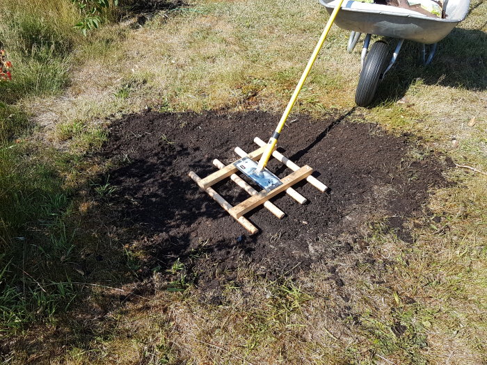 Hemmagjord dressraka av slipverktyg och kvastskaft på nyjordat markstycke, skottkärra i bakgrunden.