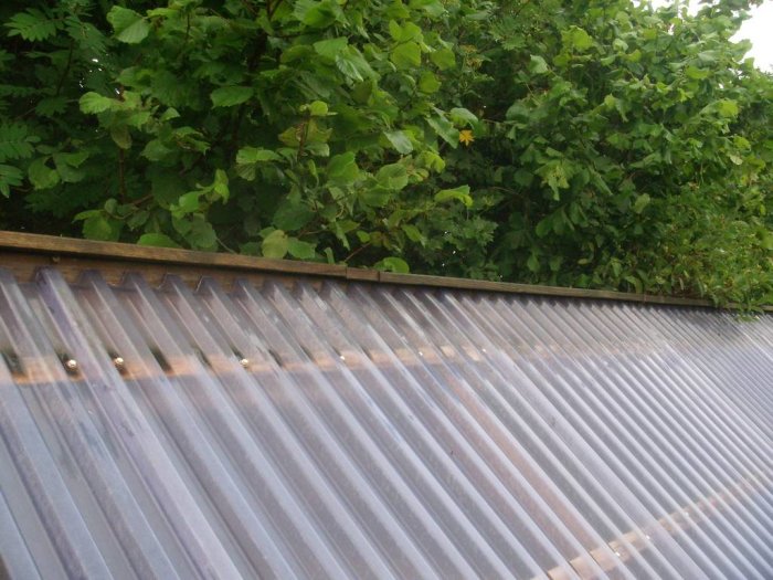 Corrugated metal roofing with foam sealant between angle plate and lower sheet, meeting a wooden edge, with greenery in background.