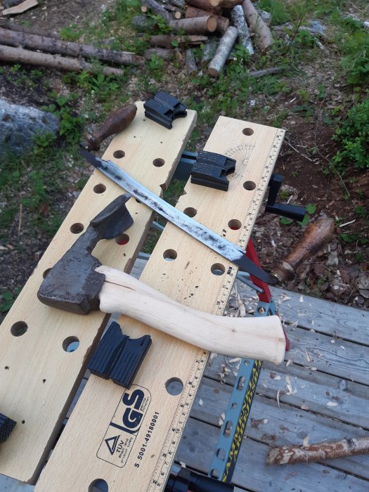 Verktygsvård med nyslipad bandkniv och avbrutet yxskaft på arbetsbänk, i bakgrunden syns skog och ved.