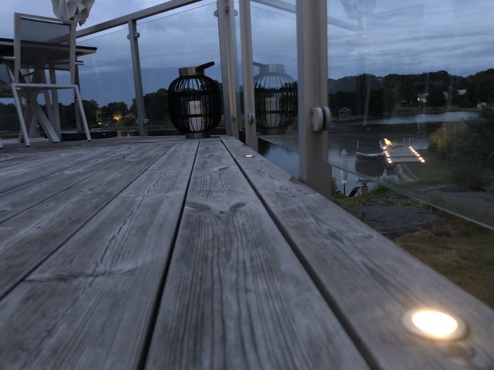 Träaltan vid skymning med vit utemöbler och dekorativa lyktor, utsikt över en sjö och belysning längs kajen.