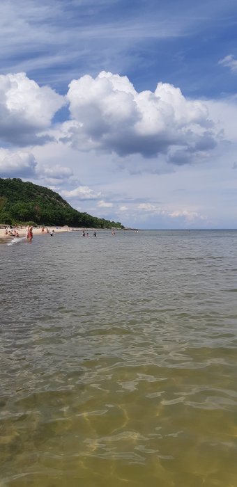 Bild av en solig strand vid klart vatten med badande människor och gröna kullar i bakgrunden.