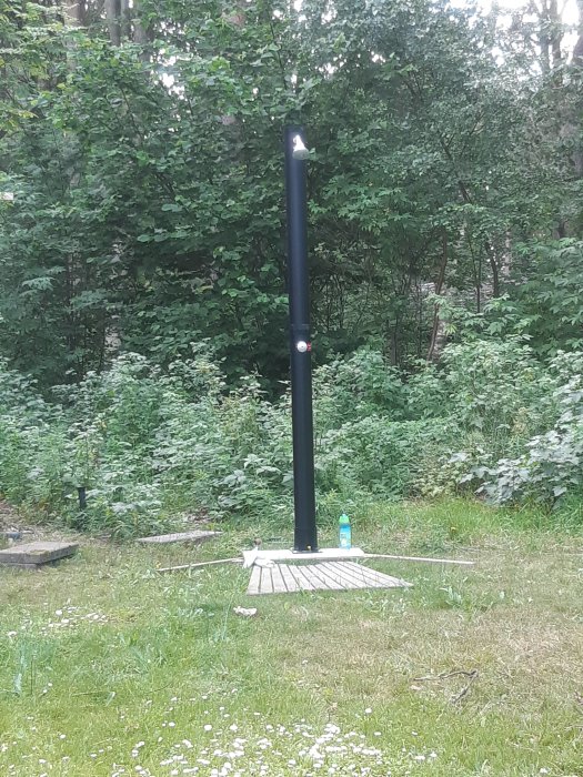 Svart utomhusdusch installerad på en träplattform i en trädgård med gröna buskar i bakgrunden.