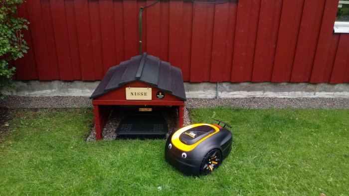 Robotgräsklippare framför sitt laddningsgarage med namnet 'Nisse' på en gräsmatta.