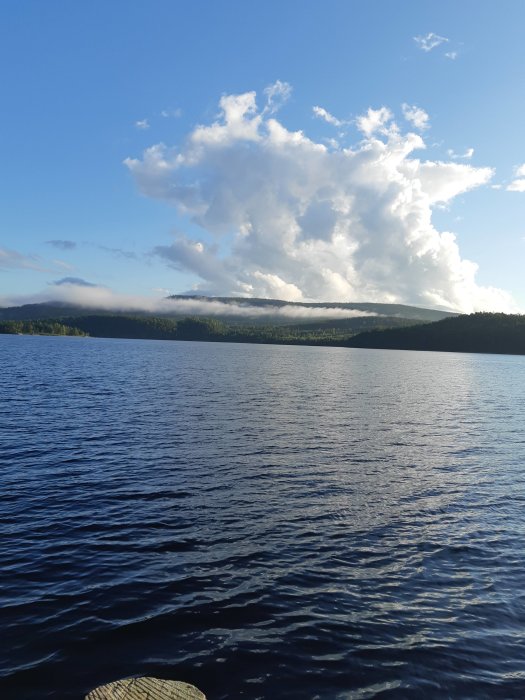 Solbelyst sjö med moln och skogklädda berg i bakgrunden.