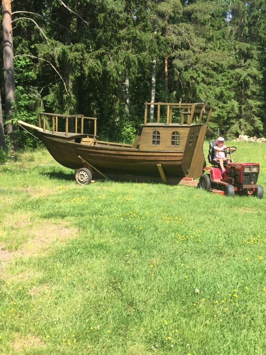 En person kör en traktor som drar en stor trälekstuga formad som ett skepp på en gräsmatta.