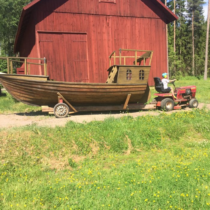 Person kör en traktor som drar ett stort träbåt på en kärra framför en röd lada.