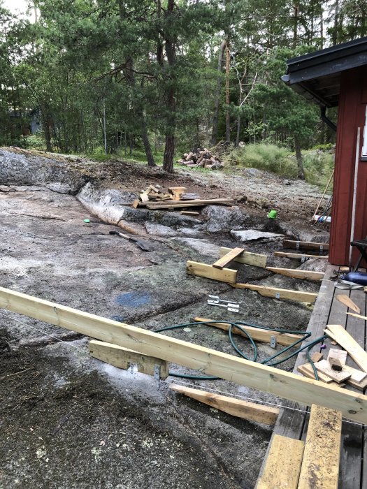 Byggplats med rensat berg och påbörjade trätrappsteg med utspridda verktyg och byggmaterial i en skoglig miljö.