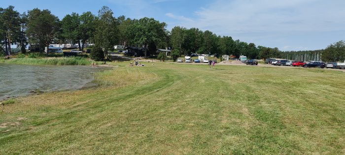 Strand vid Kalvholmens Camping med få besökare, träd, bilar och husbilar i bakgrunden.