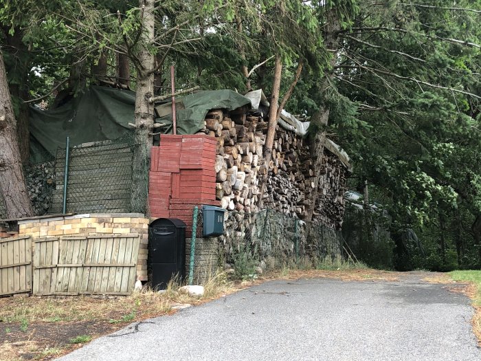 Stapel av ved i olika höjder upp till 5-6 meter med och utan presenningar vid träd och staket.