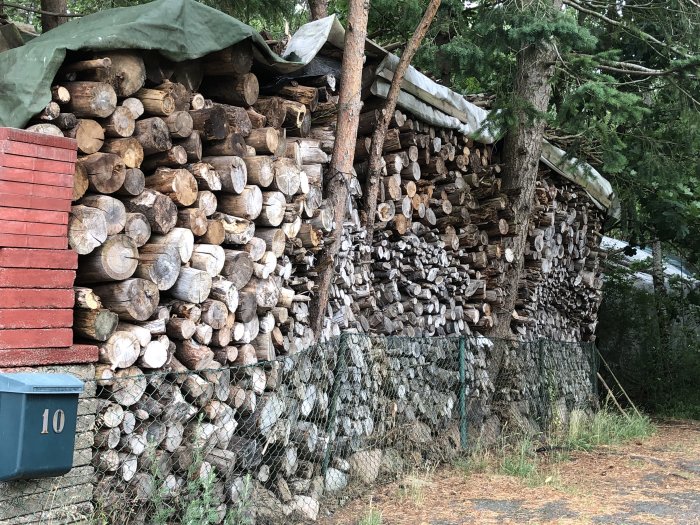 Stor vedhög framför trädfort, delvis täckt av presenning, intill rödfärgat hus och staket.