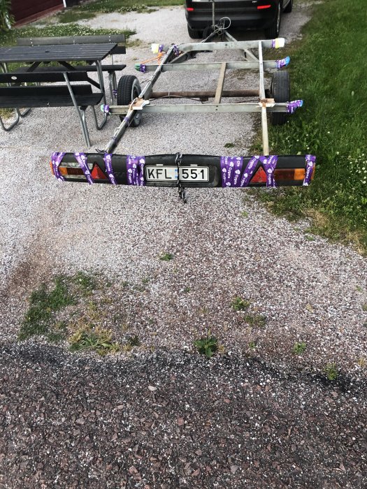 Tom släpvagn parkerad på grusunderlag med registreringsskylt synlig och bastumaterial fastsäkrat.