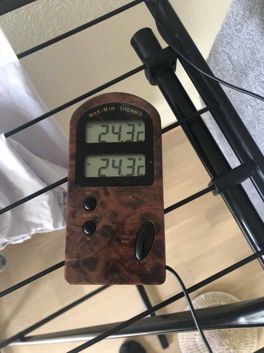 Digital termometer som visar samma temperatur inne och ute, placerad på ett bordsställ.