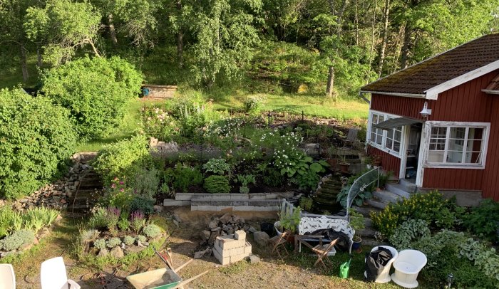 Vy över en lummig trädgård med en pågående anläggning av vatten-rabatt nära ett rött hus.