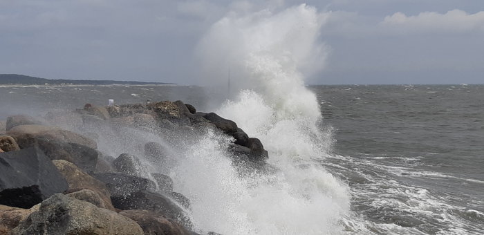 Stora vågor bryter mot klippor under storm, skapar vattensprut mot himmel.