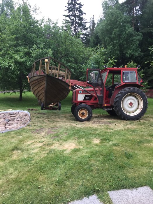 Traktor i grönt landskap som lyfter en stor träkonstruktion, med stenklädd yta och dräneringsslang synlig i förgrunden.