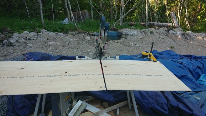 Kap-/gersåg med träskiva framför och laserlinje på skivan, placerad utomhus på ett sågbord med spån och verktyg omkring.