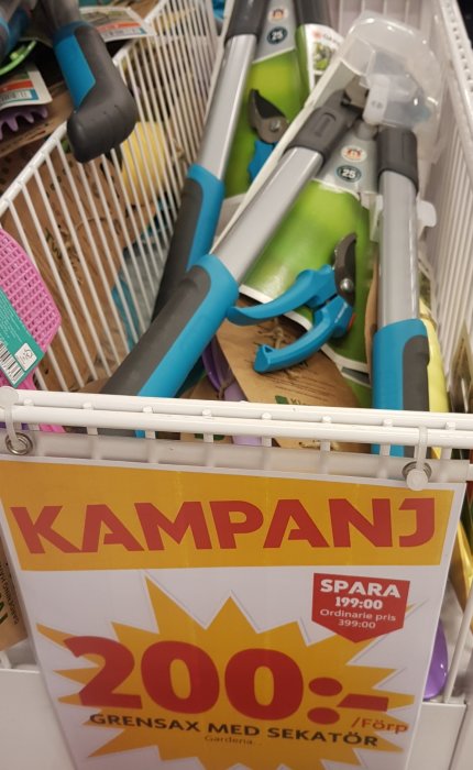 Kampanjskylt med texten "200 kr, grensax med sekatör" framför trädgårdsverktyg i en varukorg på Ica Maxi.