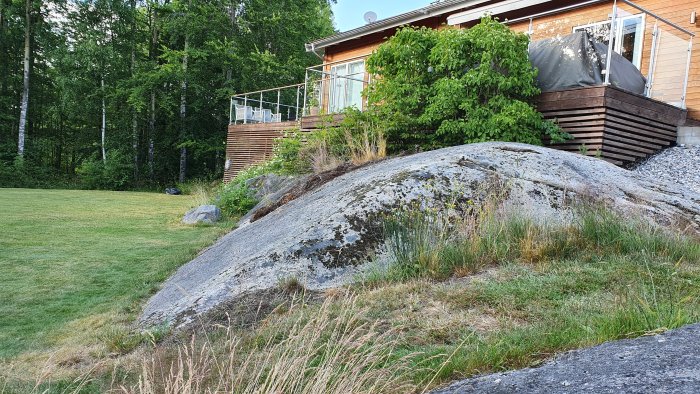 Stor grå häll på ett grönt område nära hus med träveranda och skog i bakgrunden.