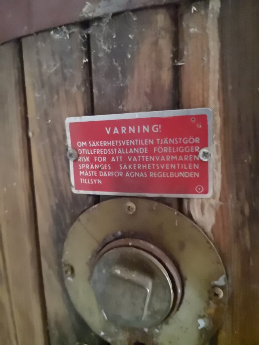 Gammal varmvattenberedare med roterbar säkerhetsventil och varningsskylt på en trävägg.