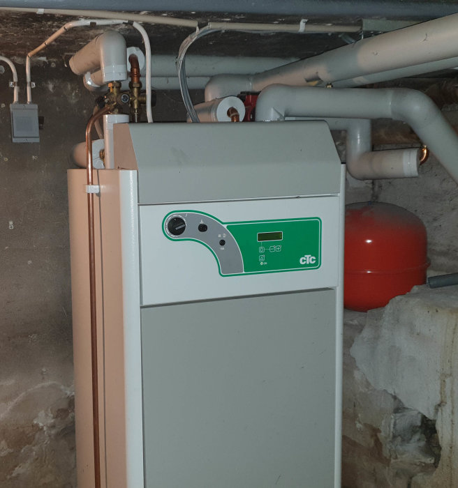 Värmepump i källare med okänd modell, logotypen CTC synlig, omgiven av rör och röd expansionskärl.