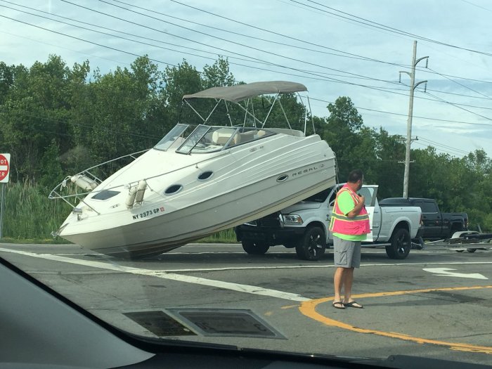 Motorbåt ovanpå släpvagn faller av och landar på väg vid korsning med förvånad person i väst.