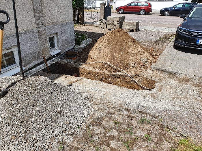 Grävd grop vid hus med synliga kabelrör nära ytan, i vägen för planerad murbyggnad.