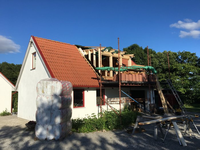 Hus under renovering med halvfärdig stomme och röd takpannor, byggmaterial och stege i förgrunden.