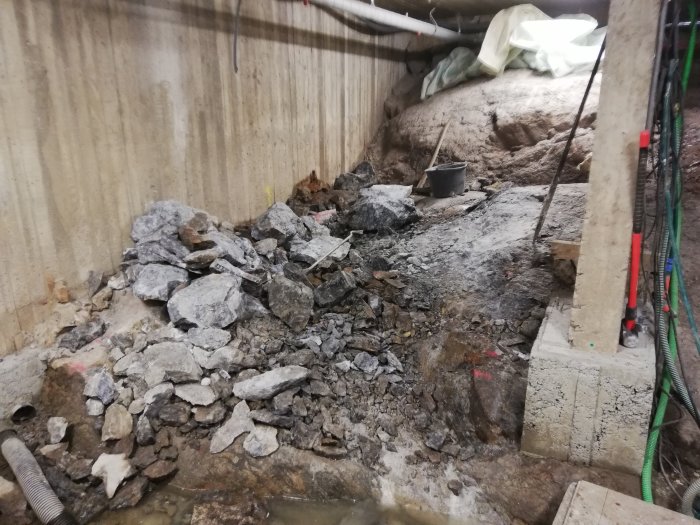 Utgrävd källare med uppbrottet sten och grus, staplade stenblock vid vägg och byggmaterial runt omkring.
