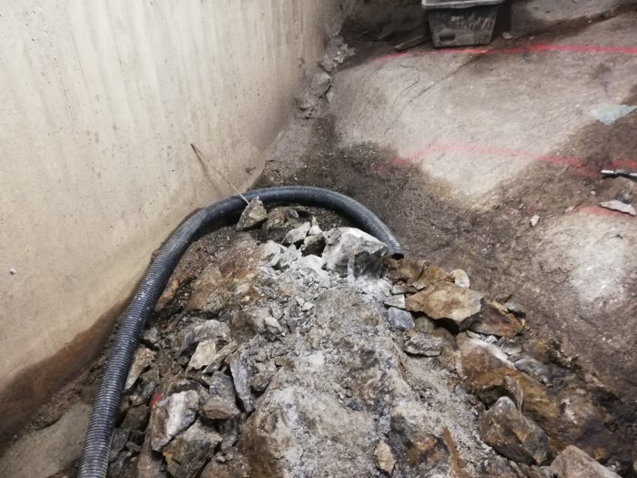 Halvkubik sten och grus med svart rör intill en betongvägg, uppmätt med röd sprayfärg.