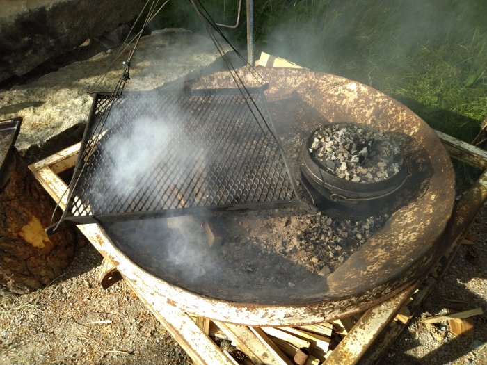 Dutch oven med glöd på lock och under, rök, vid grillplats i dagsljus.