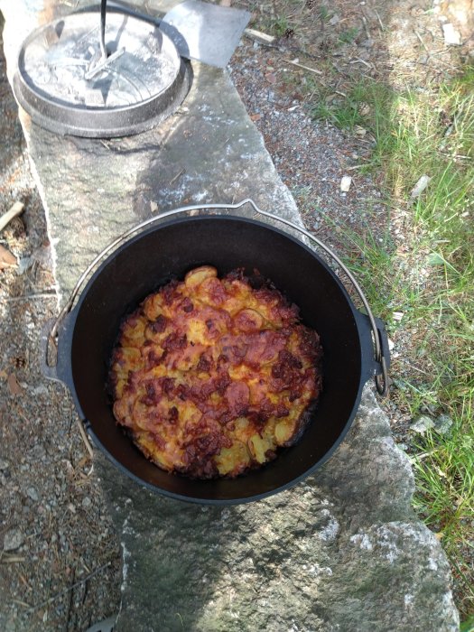 Bränd maträtt i en dutch oven på en grillplats utomhus, med lock och spade i bakgrunden.