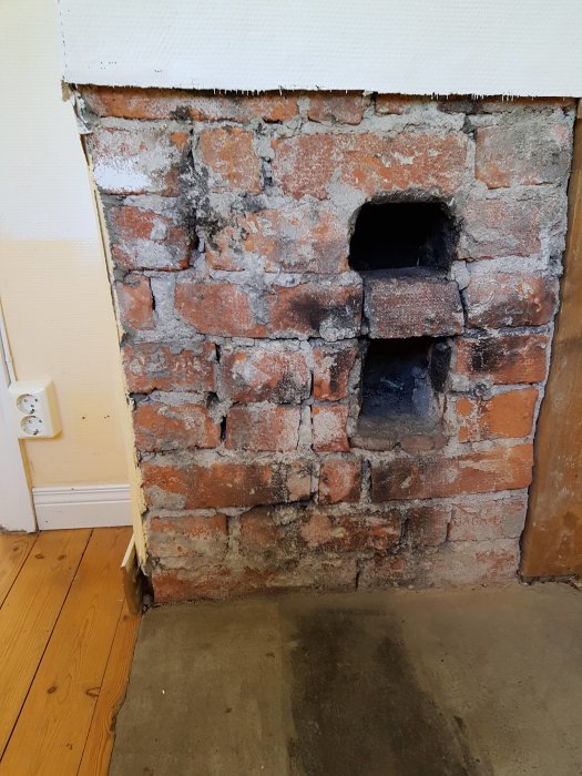 Sliten tegelvägg med tom öppning efter borttagen vedspis, omgiven av skadad puts och masonit.