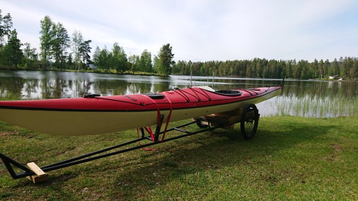 En röd och vit kajak på en hemmagjord kanotvagn nära en sjö omgiven av grönska.