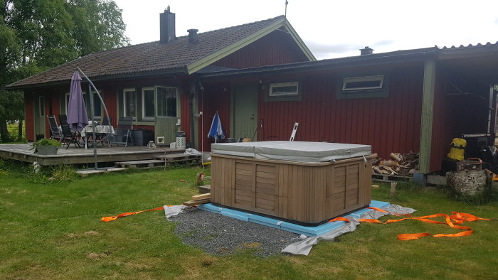 Trädäck vid hus med möbler och ett oinbyggt bad, samt planeringsmaterial synligt på gräset.