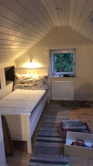 Inrett loft med snedtak, vit panel, säng, fönster, element, och öppen förvaring med leksaker på ett virkat mattgolv.