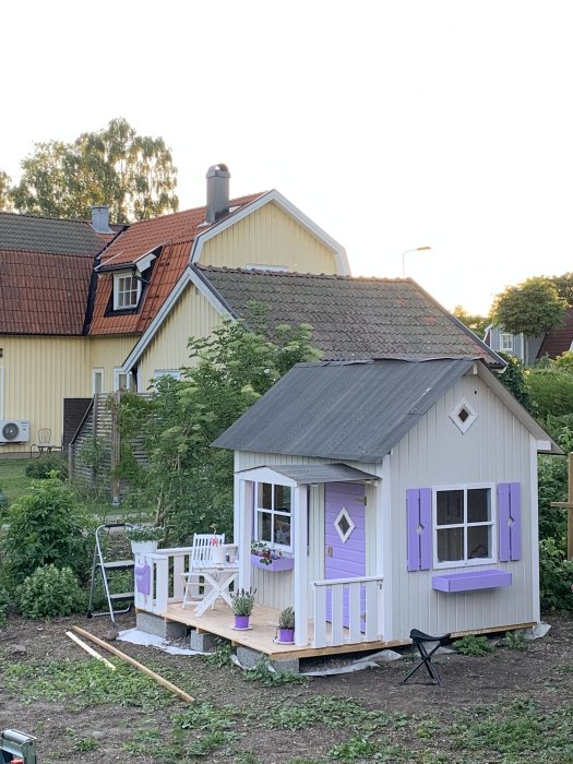 Nymålad lekstuga i ljusgrått, med lila detaljer och vit veranda, omgiven av grönska.