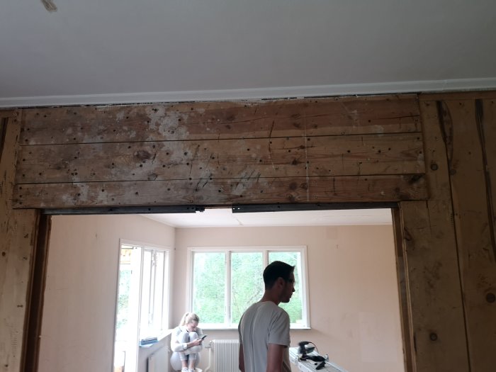 Ett rum under renovering med synligt träbjälklag ovanför en skjutdörrsöppning, två personer iakttar väggen.