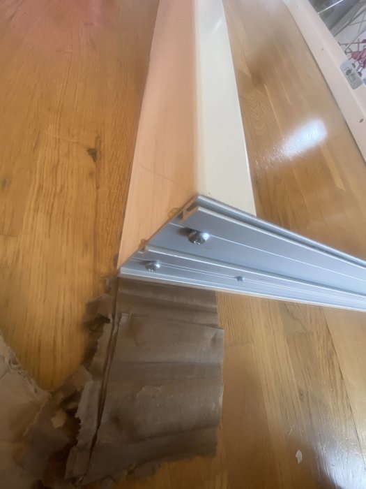 Tröskel med en omonterad bleckdetalj och synliga skruvar på ett trägolv, bredvid ett skadat kartongstycke.