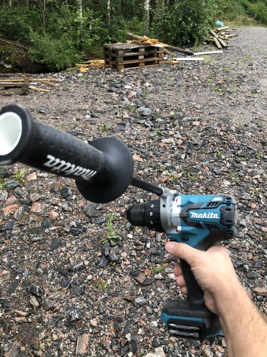 Hand håller en Makita sladdlös borrmaskin med byggmaterial och skog i bakgrunden.