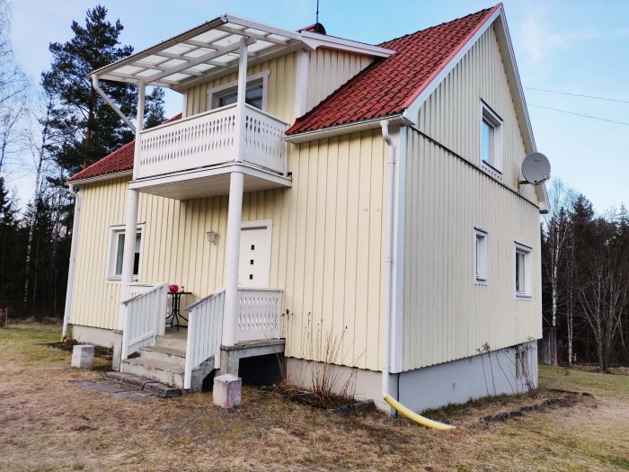 Tvåvåningshus med gult sidospår och rött tak, balkong, veranda och en satellitantenn på sidan.