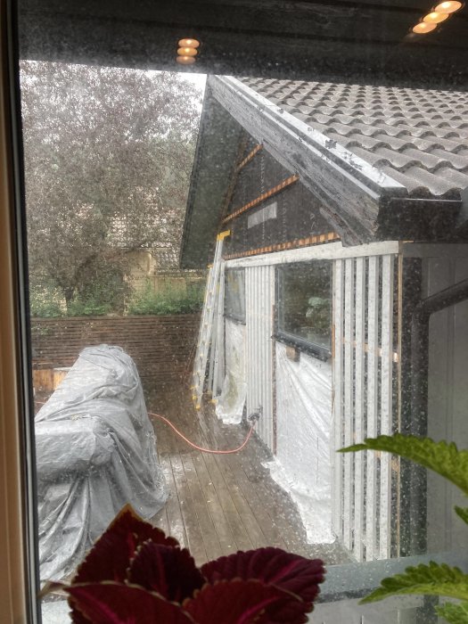Utsikt genom ett fönster på en byggarbetsplats med våt vägg och regn, täckt material på däck.
