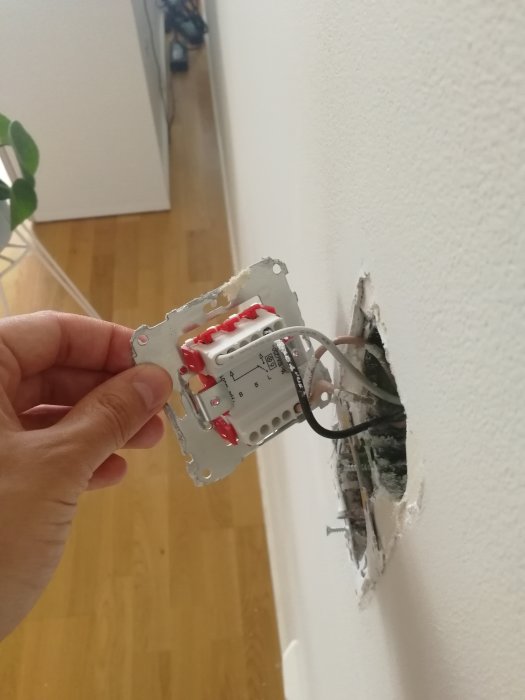 Hand håller en singel väggbrytare framför öppet eluttag i väggen, med kablar kopplade och lediga terminaler.