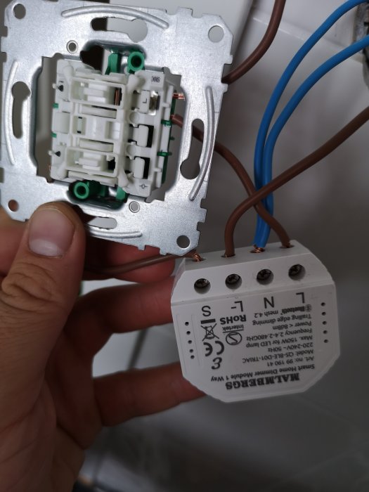 Hand håller en öppen elektrisk strömbrytare med osynliga ledningar och en fristående dimmermodul framför en vägg.