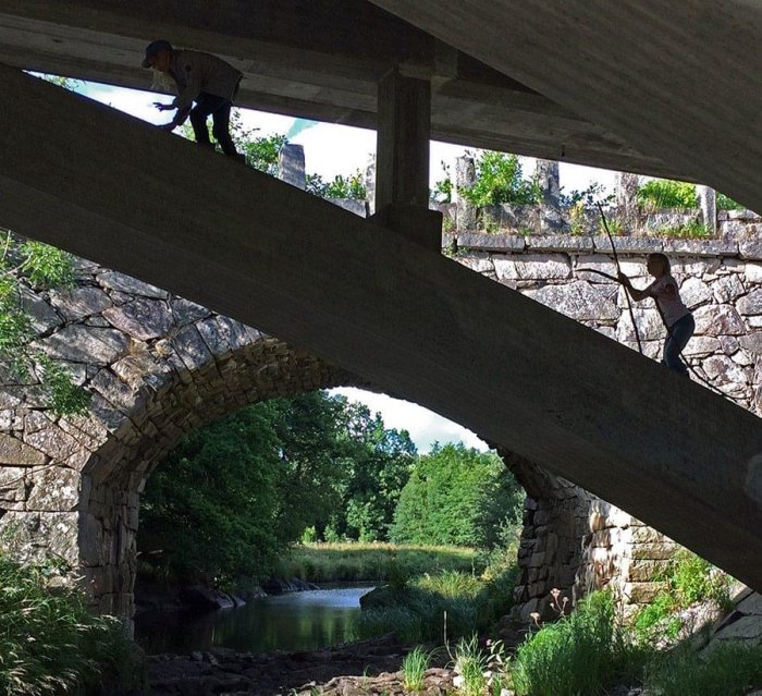 Två barn leker under en bro, en klättrar på stenvalvet och den andra balanserar på en balk.
