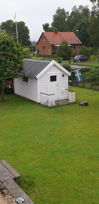 Vit friggebod med veranda och svensk flagga i en grön trädgård.