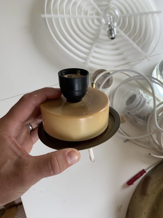 Hand håller en del av en Bumling-lampa med mässingsfot och svart lamphållare, elektriska ledningar syns ej, mot bakgrund av vägg och taklampa.