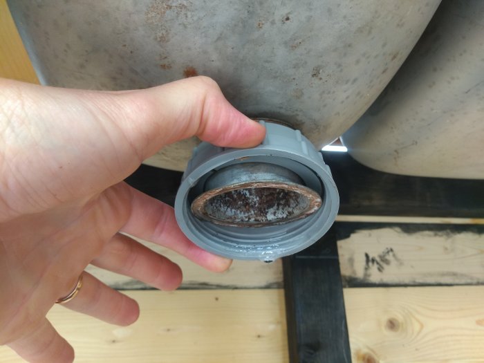 Hand håller ett vattenlås kopplat till en rostig rostfri vask över trägolv i ett byggprojekt.