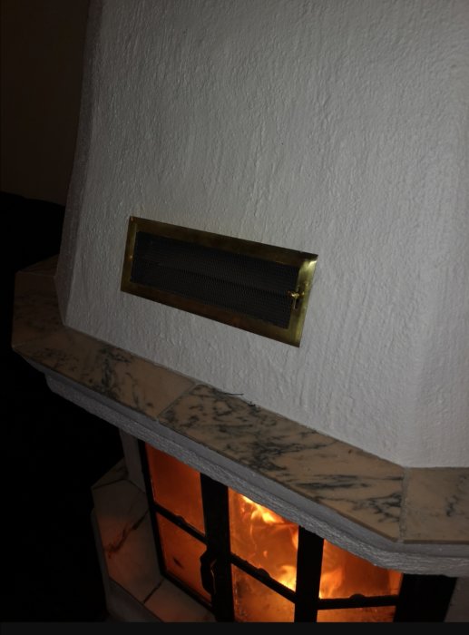 Ventilgaller ovanför brinnande kamin i öppen spis med vitputsad vägg och marmorbänk.